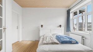 Een slaapkamer met een houten vloer, witte muren, een bed met wit- en blauwe beddengoed, een raam met blauwe gordijnen en een witte radiator.