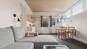 Een moderne woonkamer met een grijze bank, een klein tafeltje met een afstandsbediening, en een eethoek met stoelen.