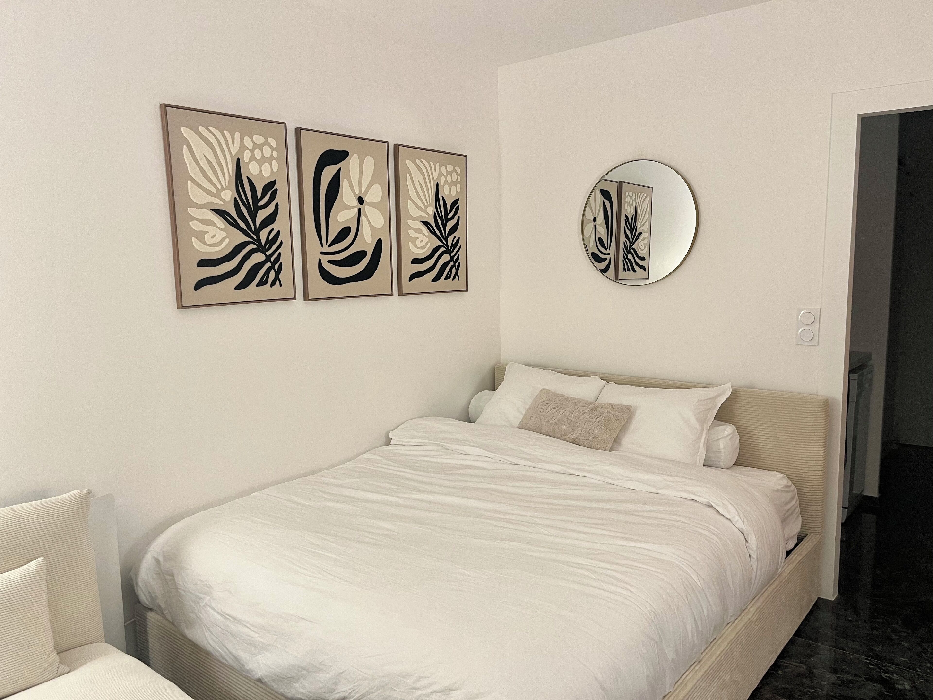 A bedroom with a bed, white bedding, a round mirror, and three abstract art pieces on the wall.