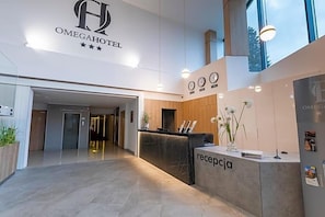 A hotel reception area with a modern design, including a reception desk labeled 'recepcja,' a clock, and a sign for 'OMEGAHOTEL.'