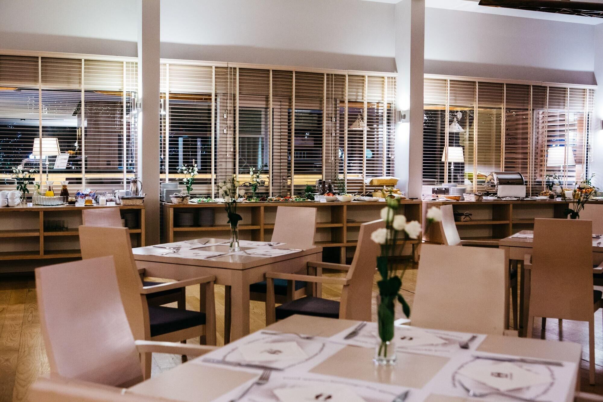 En restaurant med flere borde, stole og et stort vindue med persienner.
