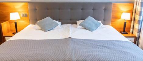 Ein Doppelbett mit grau gestopfter Bettdecke, zwei Kissen, einem gepolsterten Kopfteil und zwei Nachttischlampen.