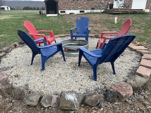 A fire pit surrounded by four Adirondack chairs, one red and three blue, set in a gravel area with a brick border.