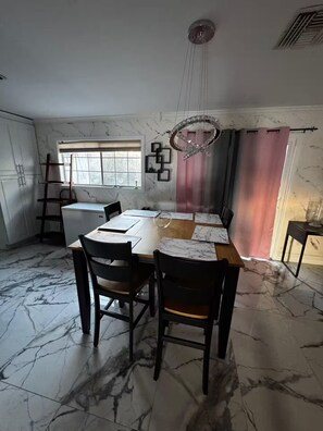 Ein Essbereich mit Marmorboden, einem Holztisch und Stühlen. Ein Fenster mit Jalousien, eine Kristallleuchter und ein pinkfarbener Vorhang sind ebenfalls zu sehen.