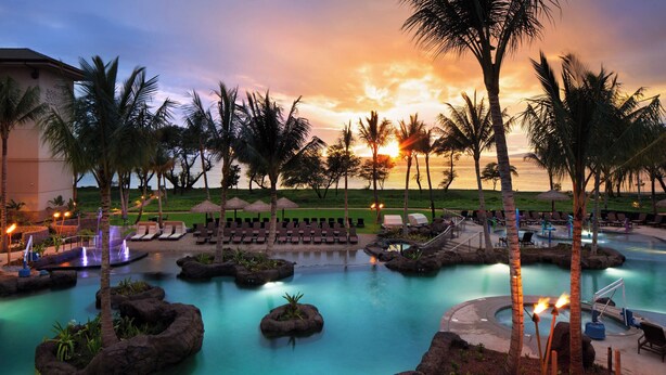 A resort pool area with lounge chairs, palm trees, and a sunset view.