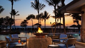 A poolside area with lounge chairs, a fire pit, and a view of the ocean at sunset.