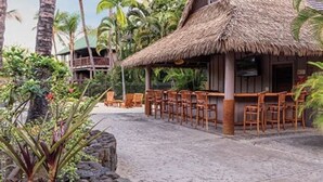 Una struttura con tetto di paglia, con sedie e tavoli di legno, circondata da vegetazione tropicale.