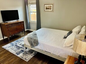 Une chambre à coucher comprenant un lit, une commode, un téléviseur, une fenêtre avec des rideaux et un tableau encadré accroché au mur.