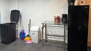 Une petite cuisine équipée d’un réfrigérateur noir, d’une machine à café et d’une étagère en verre où sont rangés divers objets. On y trouve également une poubelle, une serpillière et un balai sur le sol.