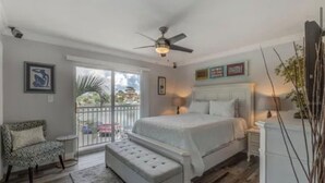 A bedroom with a bed, a bench, a nightstand, a ceiling fan, and a view of the outdoors. - Pet Friendly, St. Pete Beach Condo w/AC, 1BR+Sofa Bed, Steps to Beach! (St. Pete Beach)