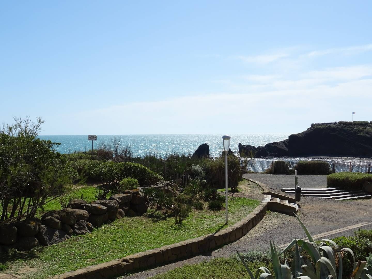 A coastal landscape with a paved walkway, rocky outcrops, and a clear view of the sea.
