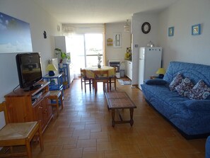 Sala de estar com televisão, móveis de madeira, sofá azul e área de cozinha com geladeira e fogão.