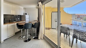 A modern kitchen with a dining area, a sliding glass door leading to a balcony, and a view of the cityscape. - CAP D'AGDE - Plage Richelieu // Réf 0168 (CAP D'AGDE)