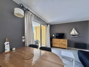 A modern living room with a wooden dining table, a TV on a wooden cabinet, and a sailboat decoration on the wall. - CAP D'AGDE - Plage Richelieu // Réf 0168 (CAP D'AGDE)