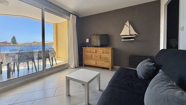 A living room with a sofa, a TV on a wooden cabinet, and a view of the sea through large windows. - CAP D'AGDE - Plage Richelieu // Réf 0168 (CAP D'AGDE)