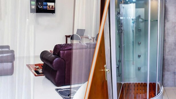 A modern hotel room with a curved glass shower enclosure, a leather sofa, and a flat-screen TV mounted on the wall. - Premium HOTEL (Douala)
