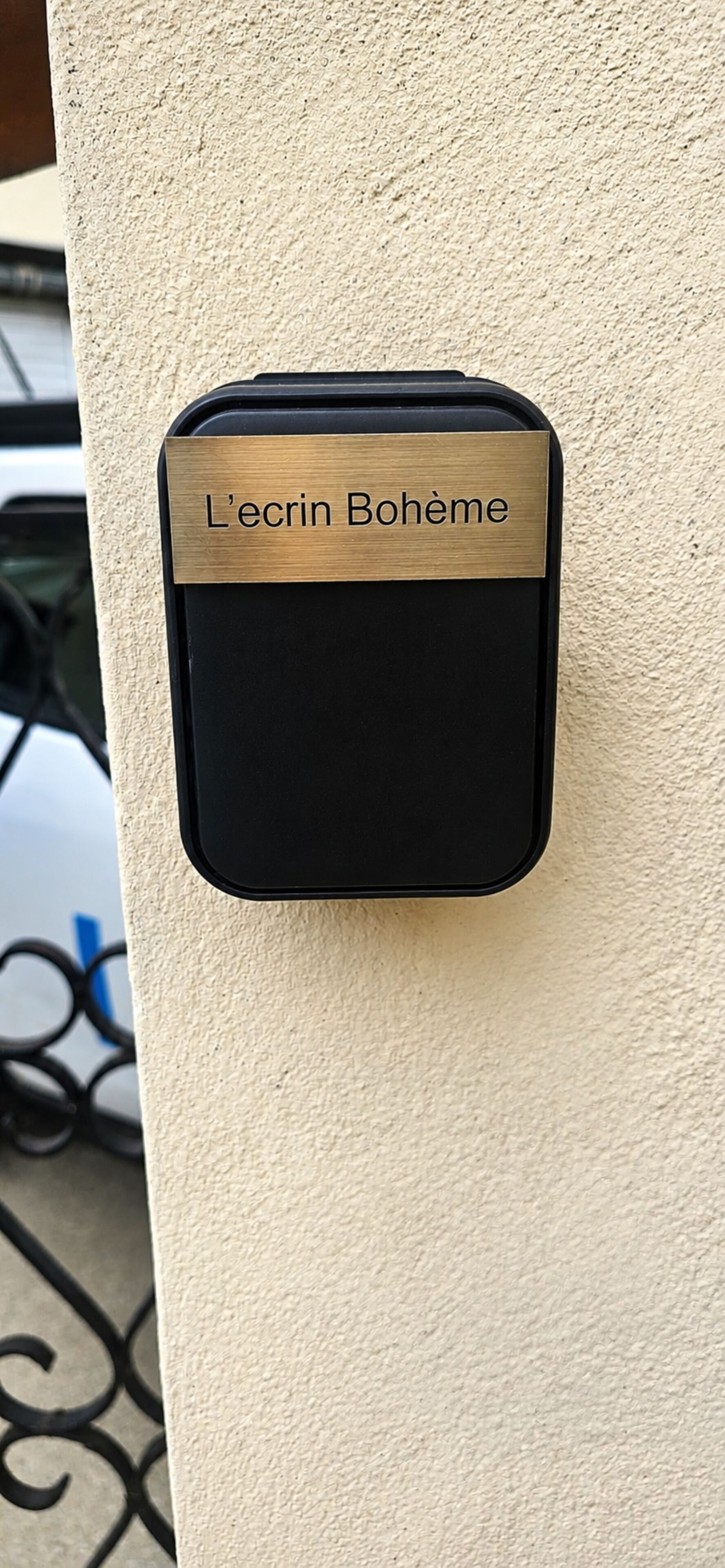 Een zwart elektronisch apparaat met een messing plaquette waarop 'L'écrin Bohème' staat, bevestigd aan een structuurwand.