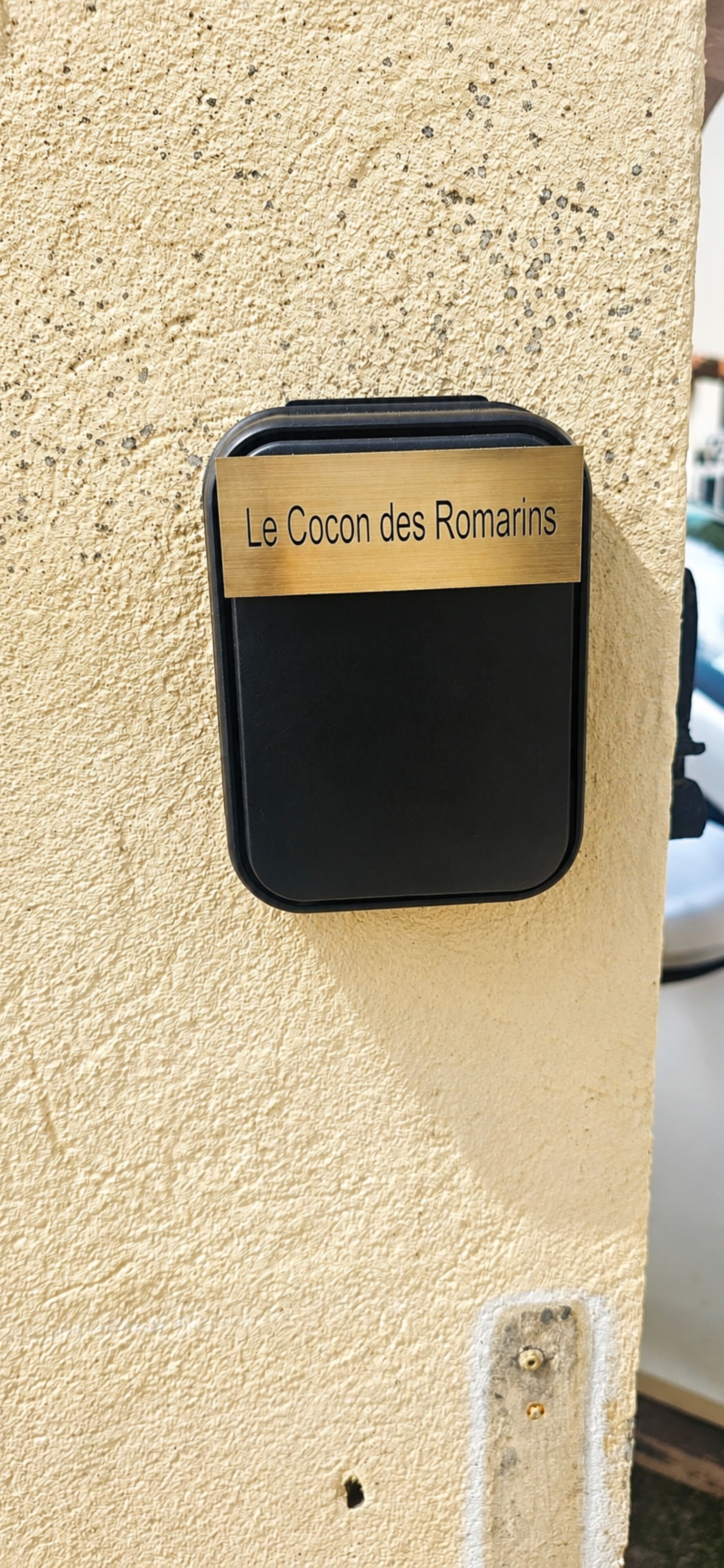 A black object with a brass plaque labeled 'Le Cocon des Romarins' attached to a textured beige wall.