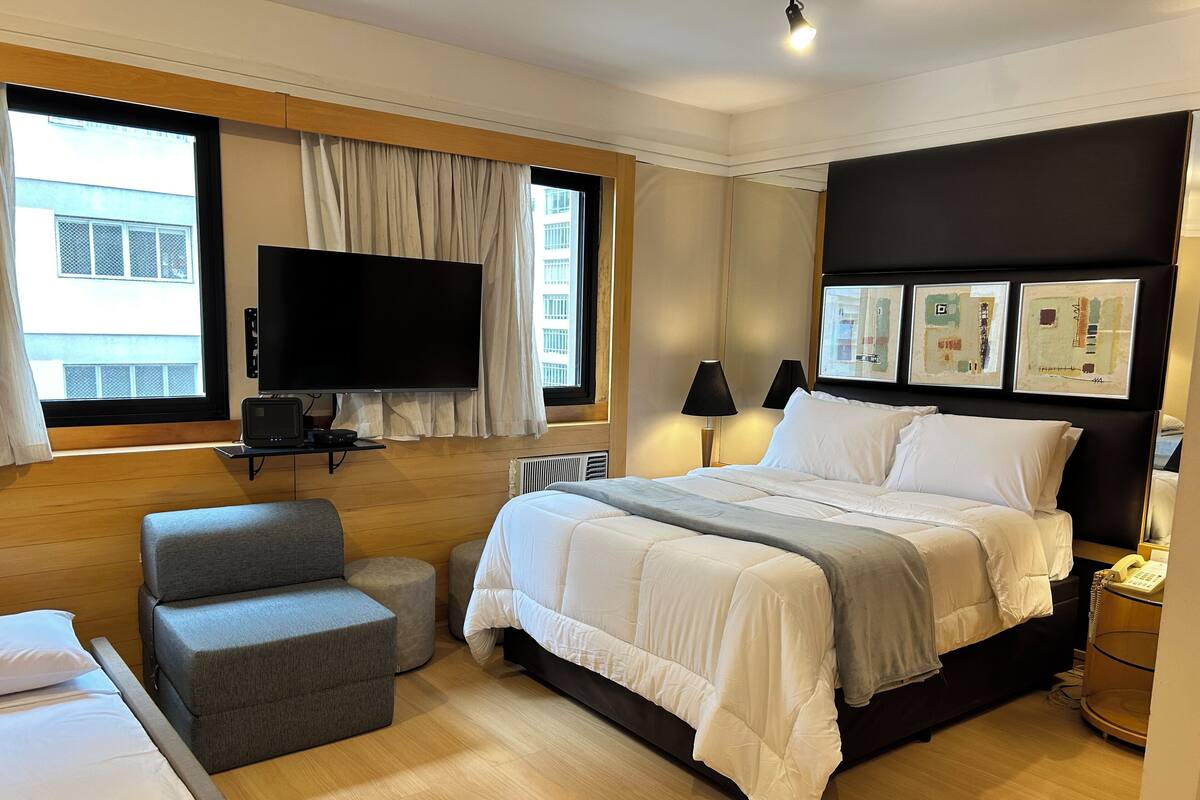Een hotelkamer met een groot bed, een aan de muur bevestigde televisie, een klein zithoekje en een raam met gordijnen.