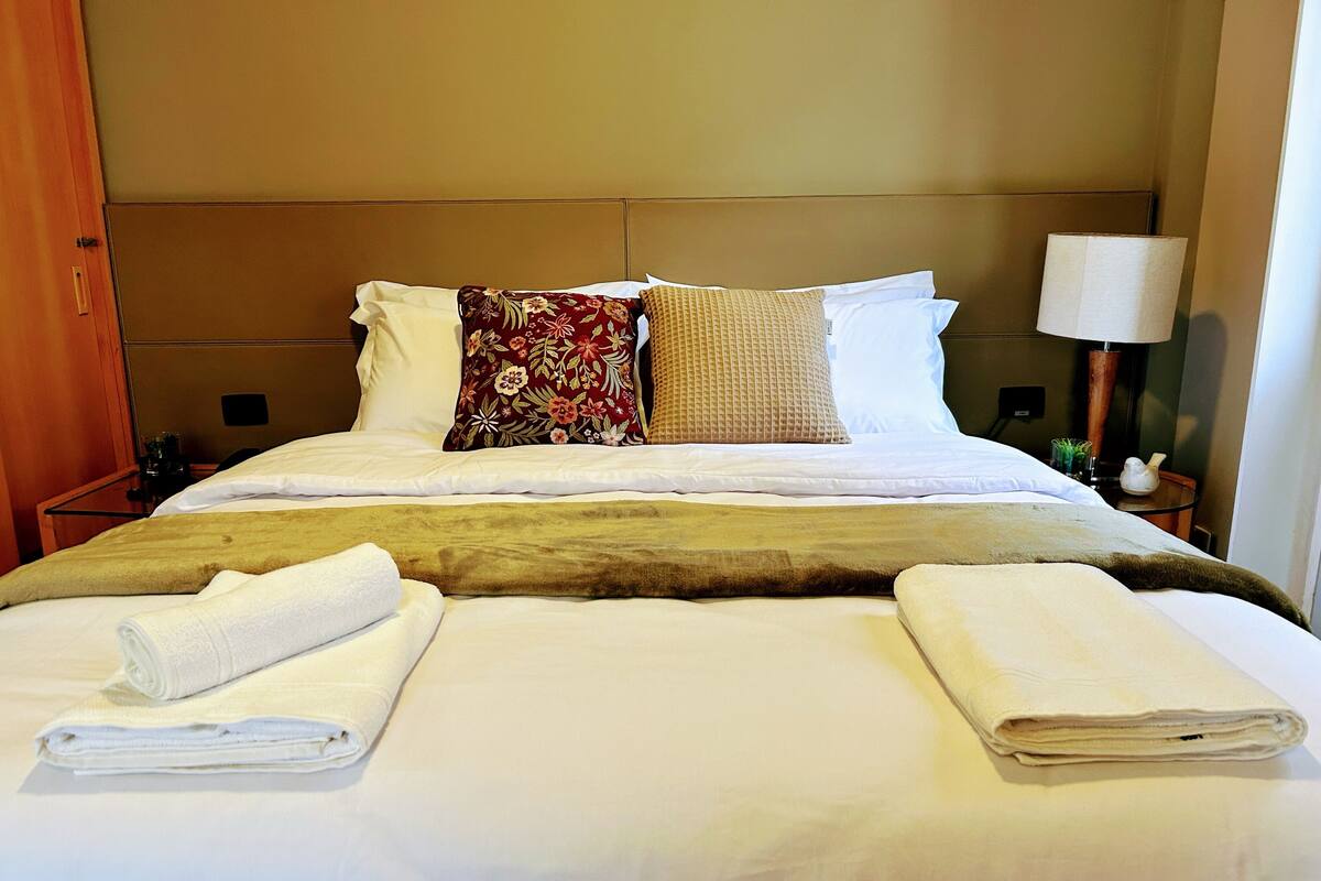 Een net opgemaakt bed met witte lakens, een bruine plaid en twee opgerolde handdoeken.