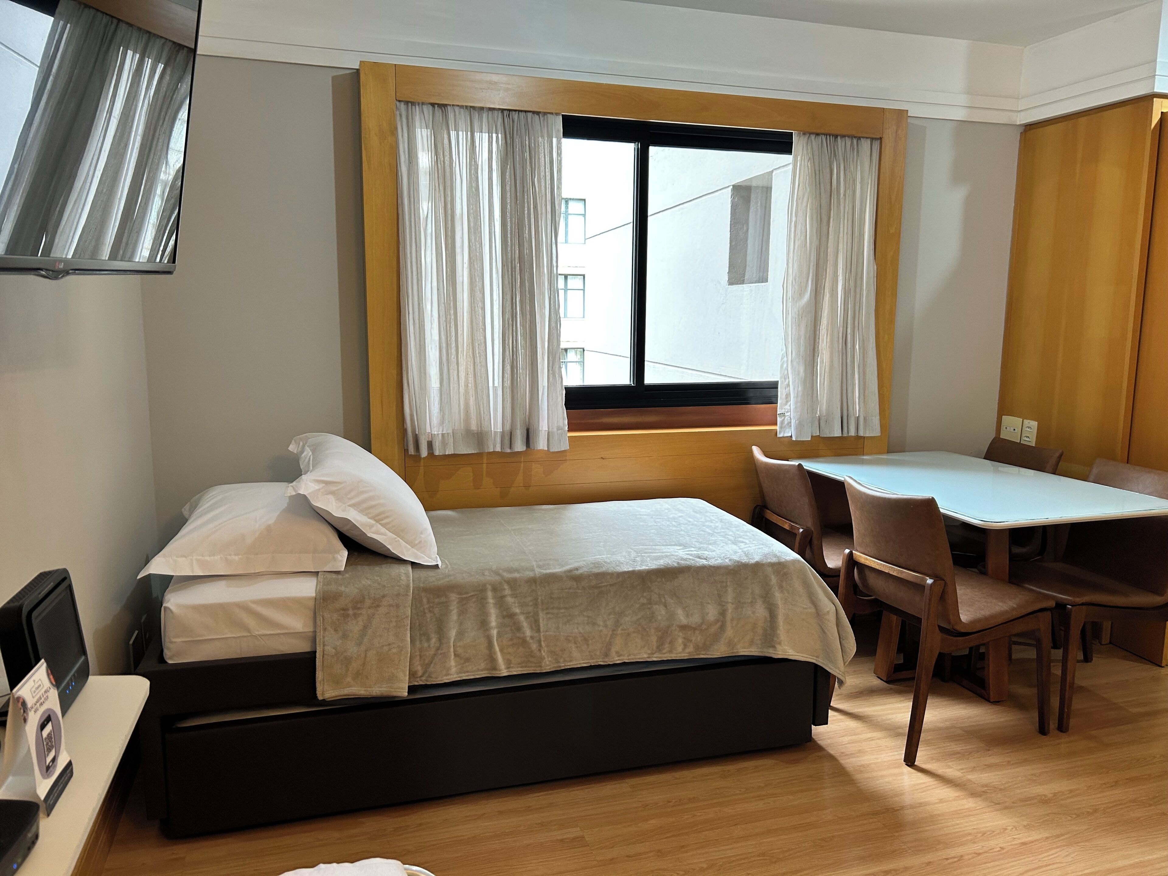 A room with a bed, a desk, chairs, a window with curtains, and a television.
