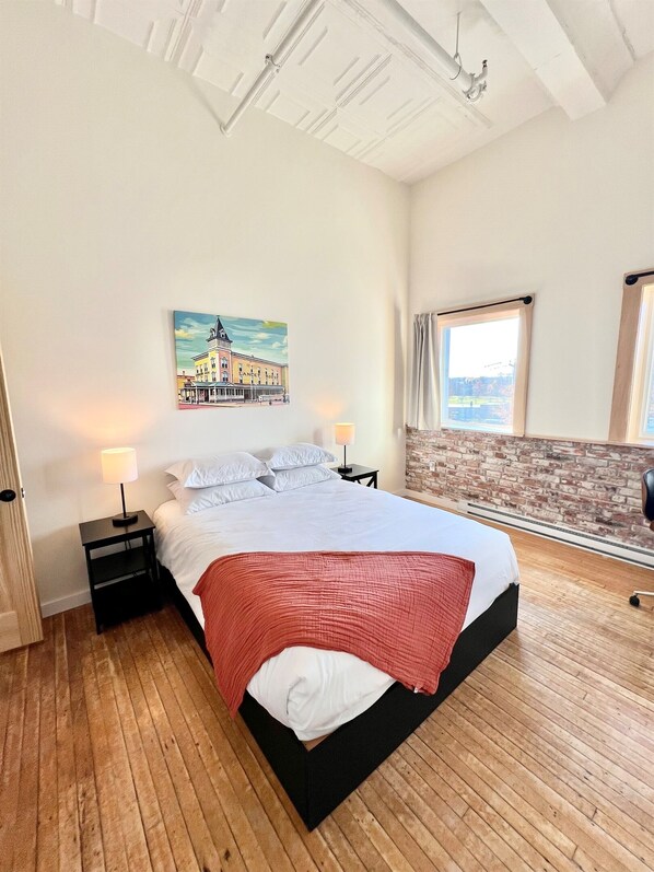 A bedroom with a large bed, a nightstand, a lamp, a painting, a window, and a brick wall.