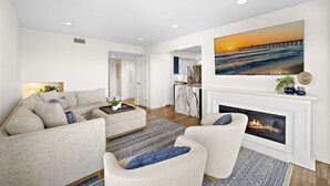 Una sala de estar moderna con chimenea, un cuadro grande de un atardecer sobre un muelle y una cocina al fondo.