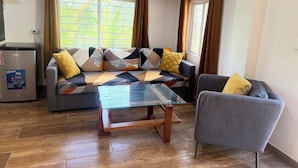 Ein Wohnzimmer mit einer Couch, einem Glastisch, einem Sessel, einem Kühlschrank und einem Fenster mit Vorhängen.