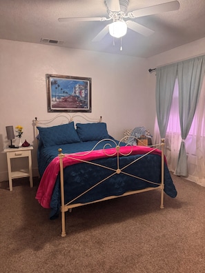 A bedroom with a bed, a nightstand, a ceiling fan, a painting, and a window with curtains.