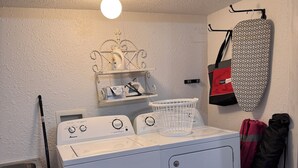 Een wasserette met een witte wasmachine en droger, een plank met schoonmaakmiddelen, een opbergrek voor een strijkplank, en een bord met de tekst: 24-uurs zelfservice wasserij.