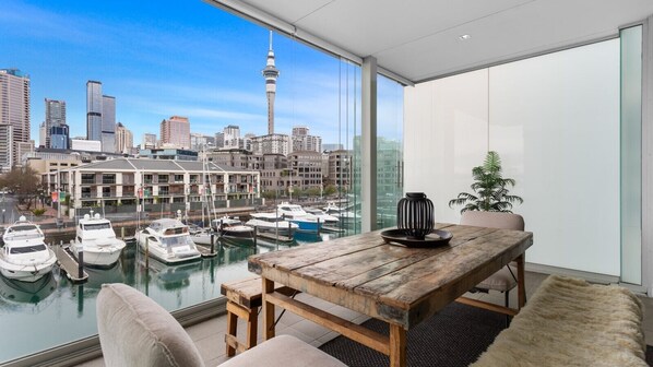 A modern interior with a wooden table and chairs, a view of a marina with boats, and a city skyline in the background. - Waterfront Marina Retreat – Pool, Gym & Parking (Auckland City)