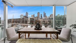 A modern dining area with a city view, a wooden table, two chairs, and a decorative vase. - Waterfront Marina Retreat – Pool, Gym & Parking (Auckland City)