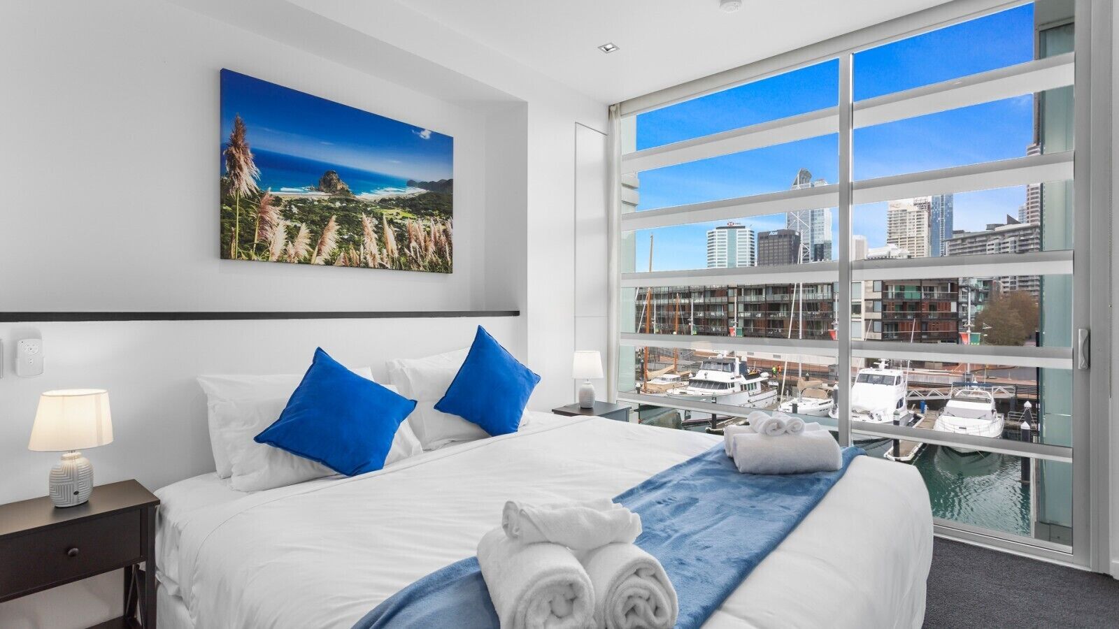 A hotel room with a large bed, a view of a marina, and a painting on the wall.