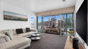 A modern living room with a sofa, two side tables, a bench, and a large window with a city view. - Waterfront Marina Retreat – Pool, Gym & Parking (Auckland City)