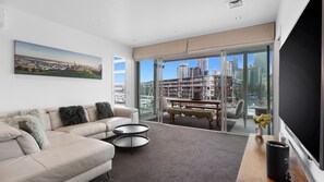 A modern living room with a sofa, two side tables, a bench, and a large window with a city view. - Waterfront Marina Retreat – Pool, Gym & Parking (Auckland City)