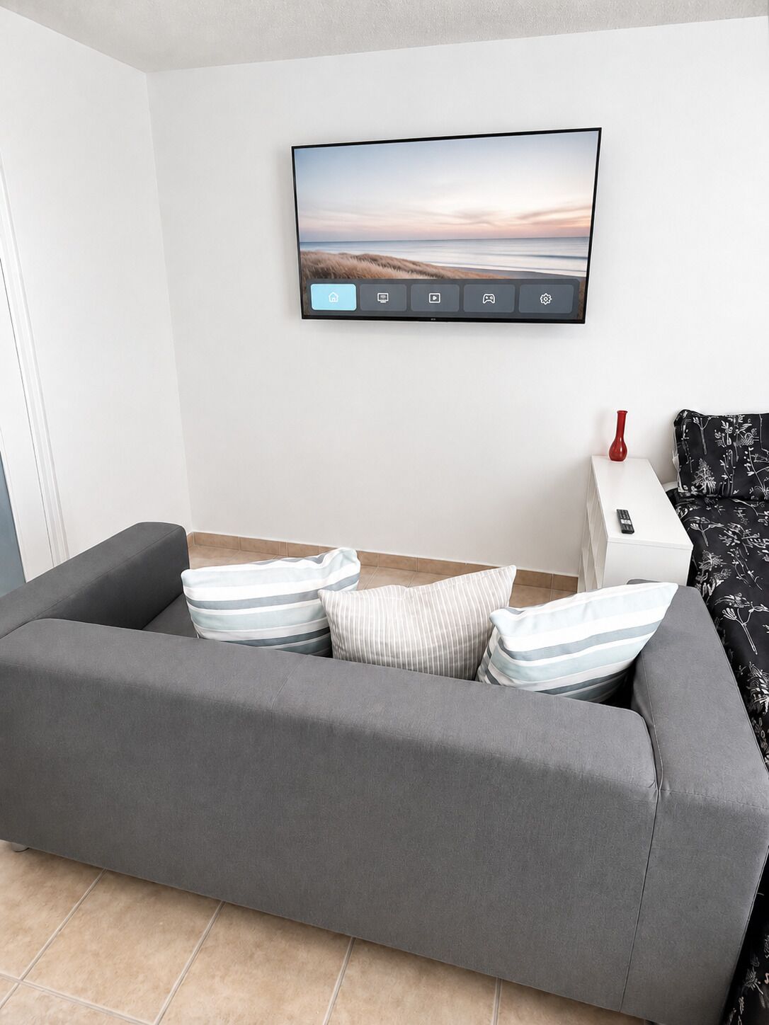 En moderne stue med en grå sofa, et fladskærms-TV monteret på væggen, et lille hvidt sofabord med en rød vase og et sort-hvidt blomstret plaide.