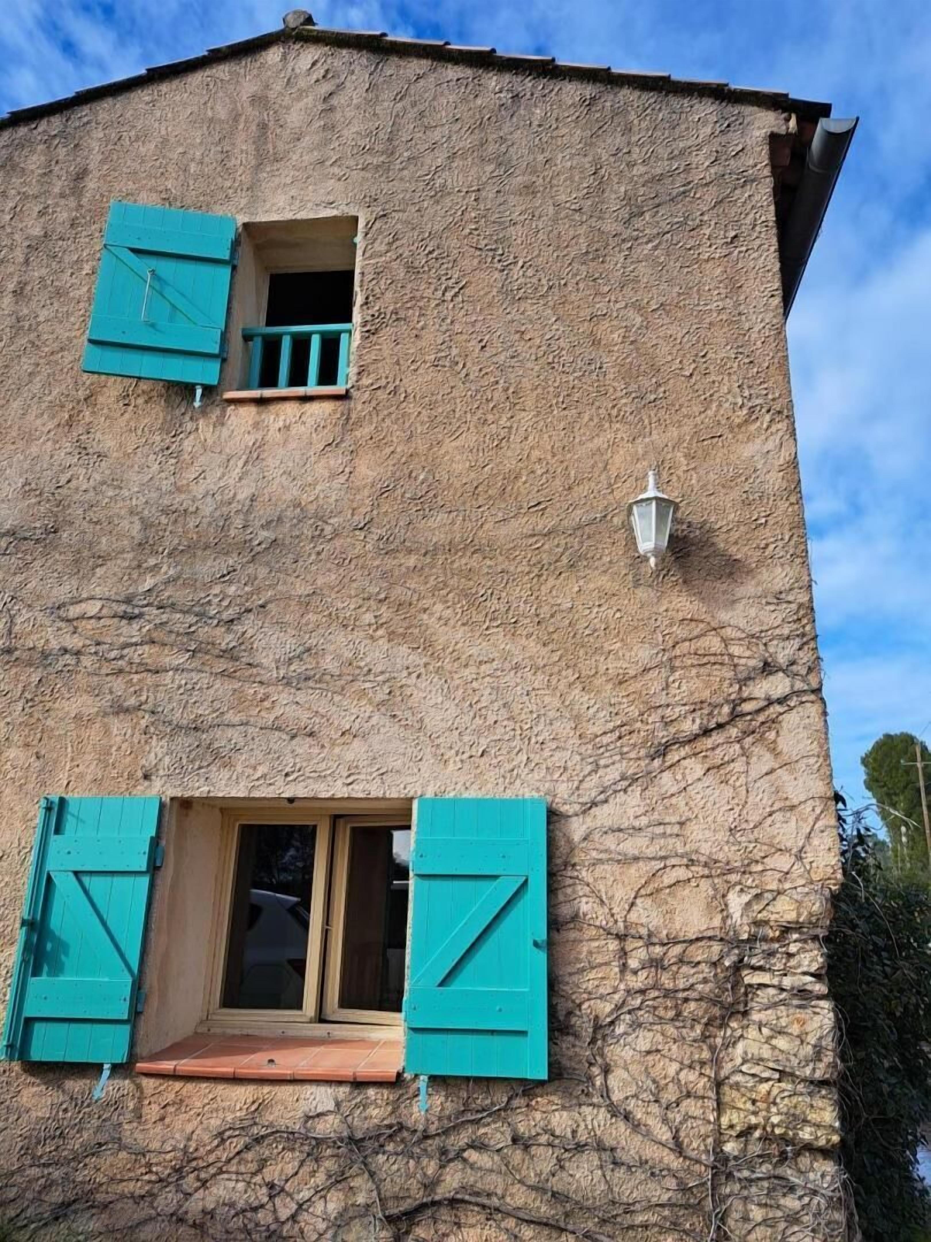 Een gebouw met een structuurrijk gevel, twee ramen met turquoise luiken en een klein, aan de muur bevestigd lampje.