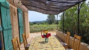 Un área de comedor al aire libre con una mesa preparada para cuatro personas, rodeada de sillas, un jarrón decorativo con flores y vistas al campo.