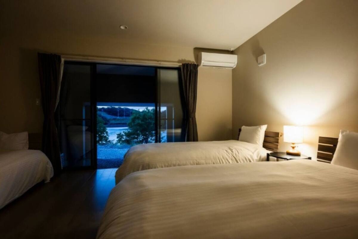 A hotel room with two beds, a window with curtains, an air conditioning unit, and a nightstand with a lamp.