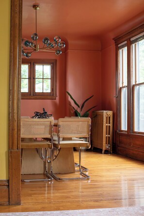 Una habitación con suelo de madera, un radiador, dos sillas de mimbre, una mesa y una ventana con vistas.