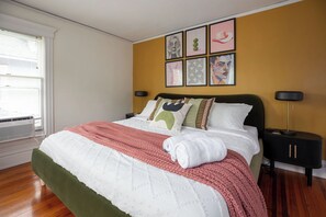 Una cama bien hecha con un cabecero tapizado en verde, un colcha blanca y una manta rosa. Hay dos mesitas de noche con lámparas negras y una pared decorada con cuadros.