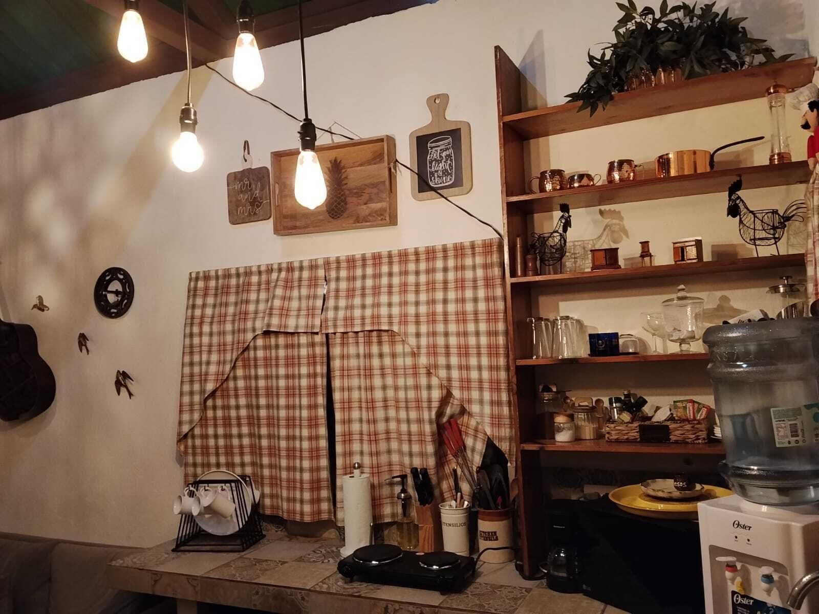 Ett kök med ett rutigt tyg som hänger som gardin, hängande lampor och hyllor fyllda med köksredskap.