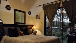A bedroom with a bed, a nightstand, a lamp, a teddy bear, and a window with curtains. - LAS AVES (Juayua)