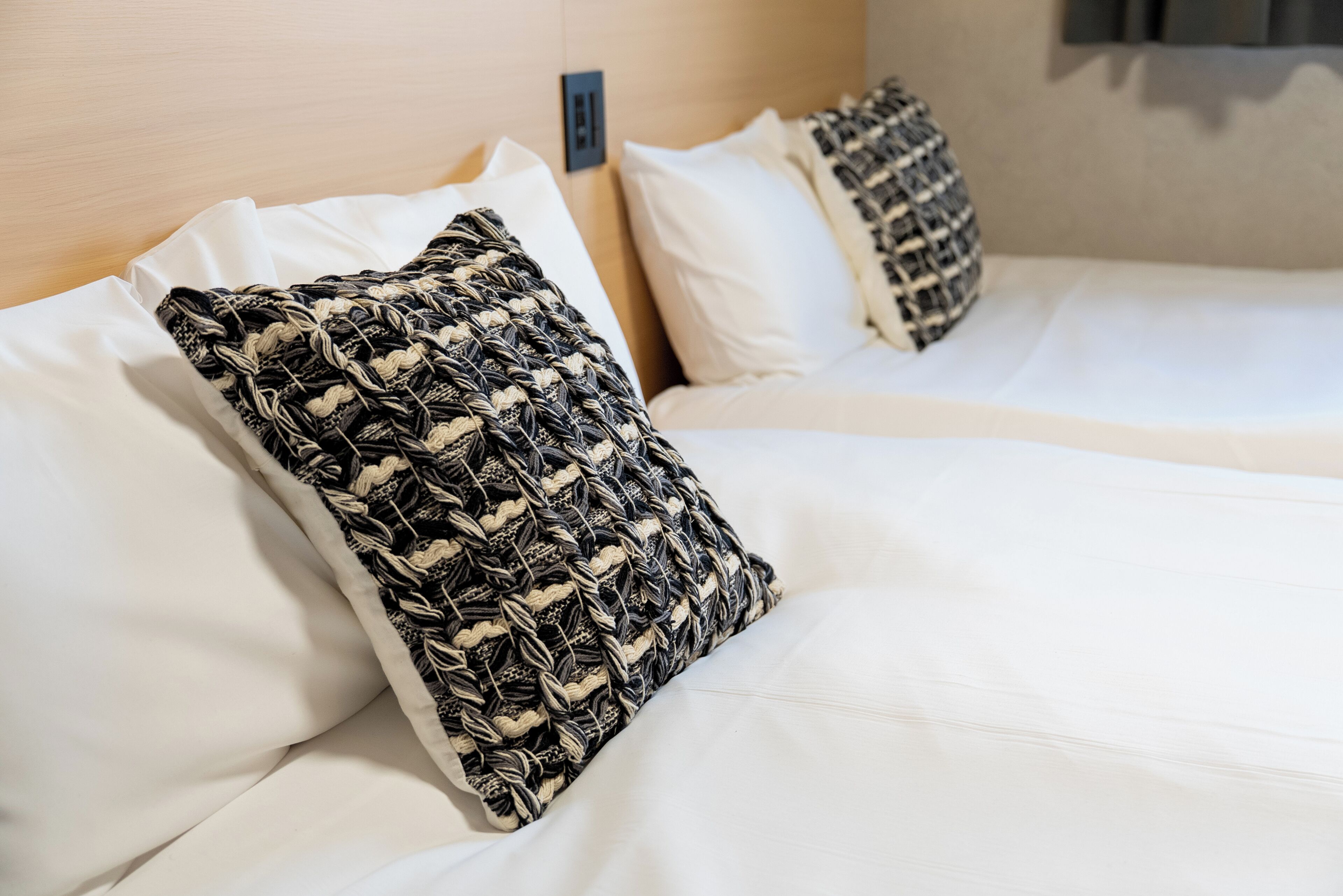 A hotel room with a neatly made bed, white bedding, and a woven-patterned pillow.