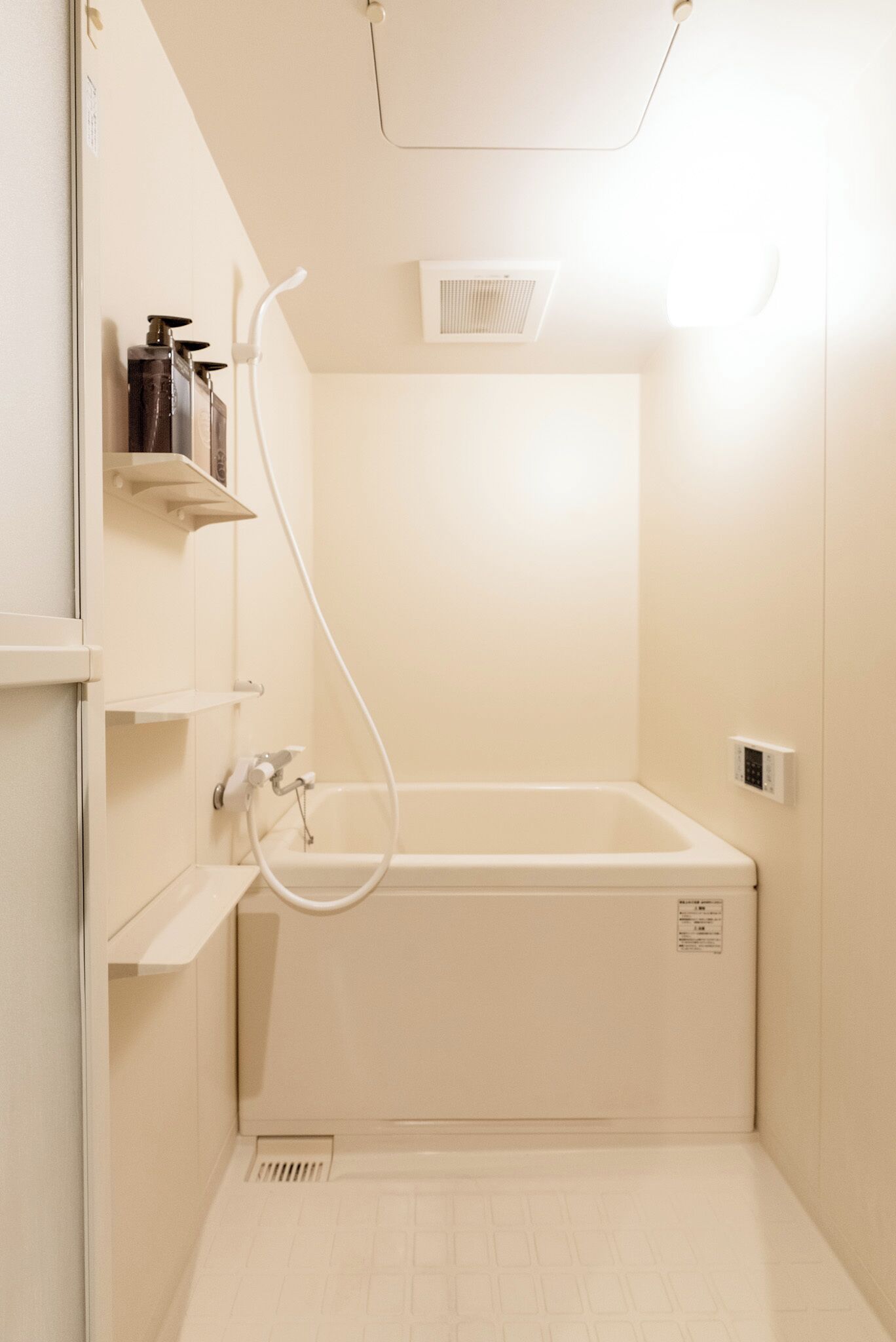 Une petite salle de bain moderne, équipée d’une baignoire, d’une douche et de toilettes. On y trouve des étagères avec des produits de toilette et une bouche d’aération au mur.
