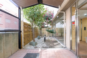Entrada externa com galeria coberta, cerca de bambu e um pequeno jardim com árvores e pedras.