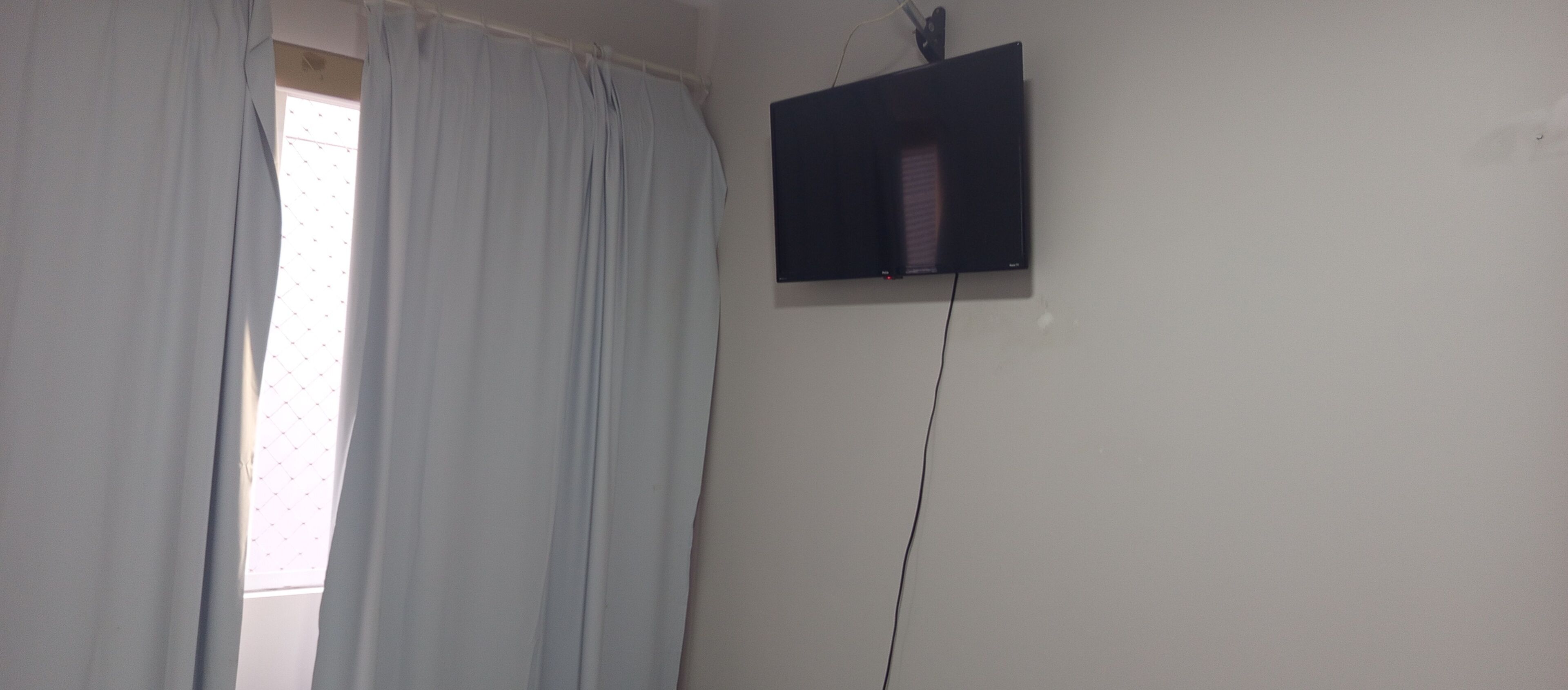 Une pièce avec une fenêtre ornée de rideaux bleu clair, un téléviseur à écran plat fixé au mur et un câble d’alimentation visible.