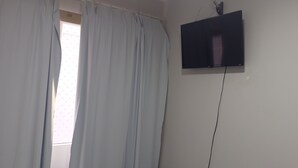 A room with a window covered by light blue curtains, a mounted flat-screen TV on the wall, and a visible power cord. - Fully furnished apartment (Rio do Sul)