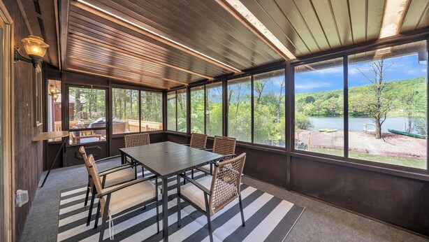 A covered patio with a dining table and chairs, large windows offering a view of a lake and trees. - New to VRBO! Lakefront I Movie Theater I Putting Green I Game Room I Bocce ball! (White Haven)