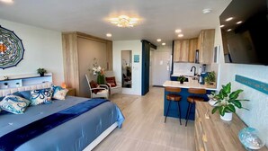 Une chambre d’hôtel moderne dotée d’un grand lit, d’un coin salon avec un fauteuil et une petite table, d’une kitchenette et d’une salle de bain visible à travers une porte ouverte.